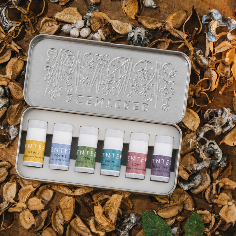 A metal tin of five essential oils by Scentered is displayed on a rustic wooden surface surrounded by dried leaves, candles, and berries. The tin is open, showing the five small blends with different favor balm.
