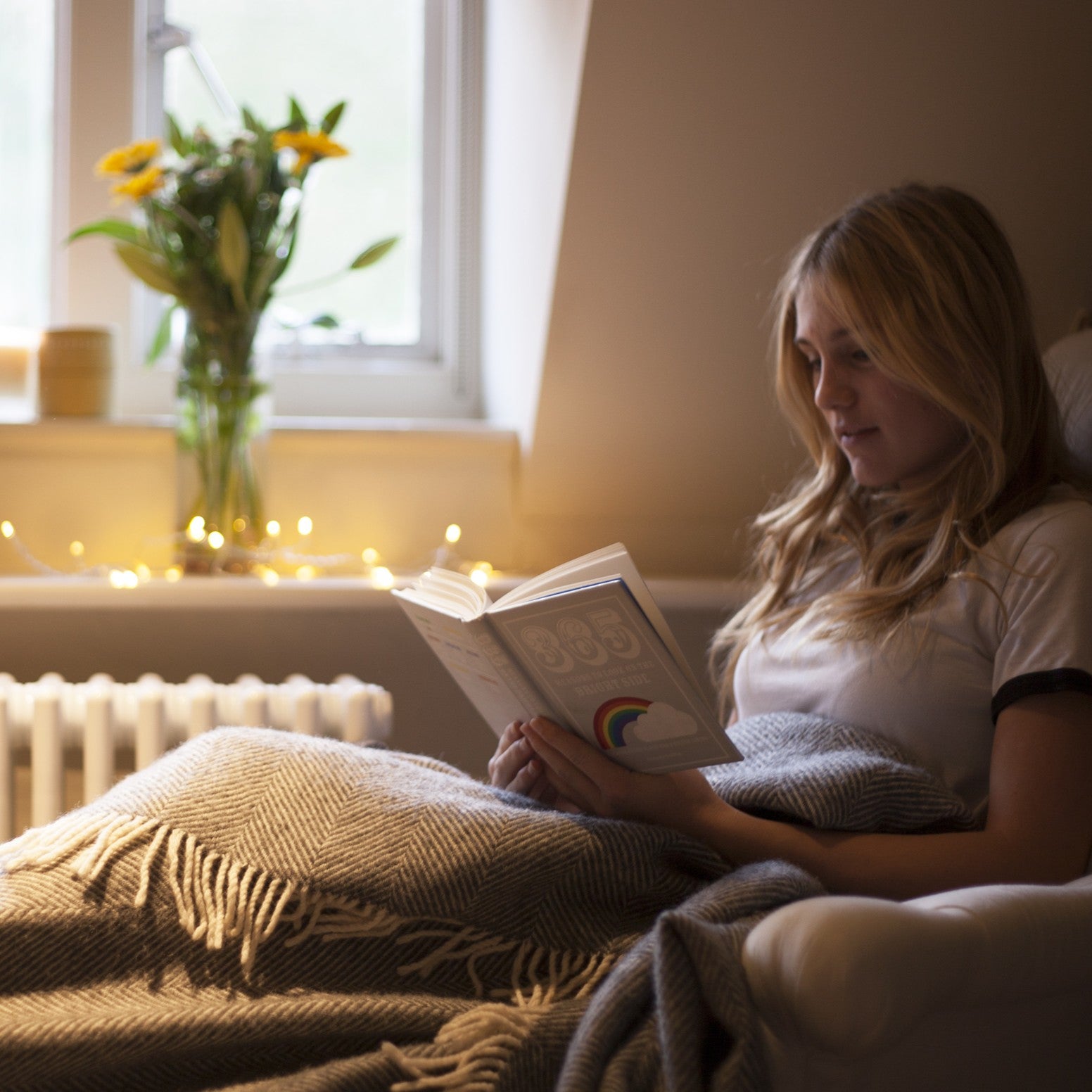 Woman reading a book in a cozy room with flowers and lights.