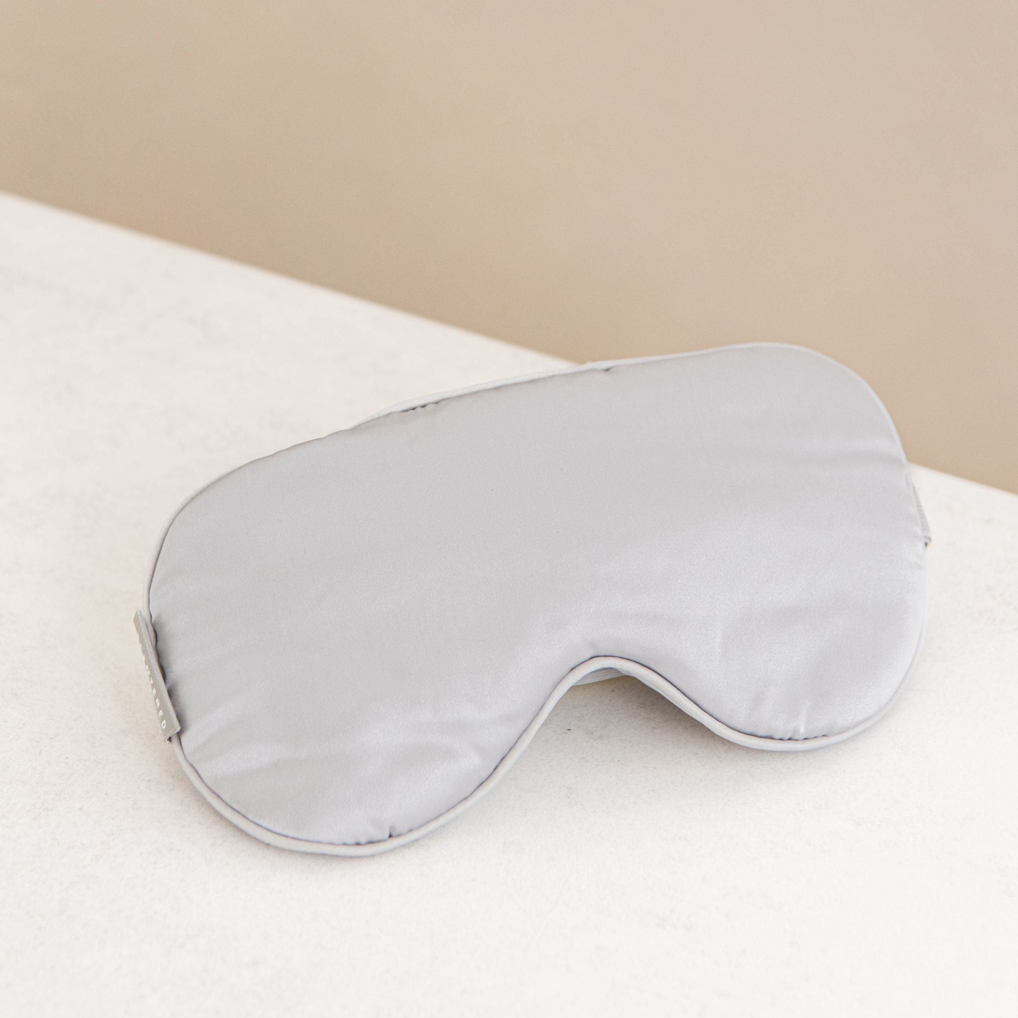 Silver silk sleep mask on a white surface with a beige background