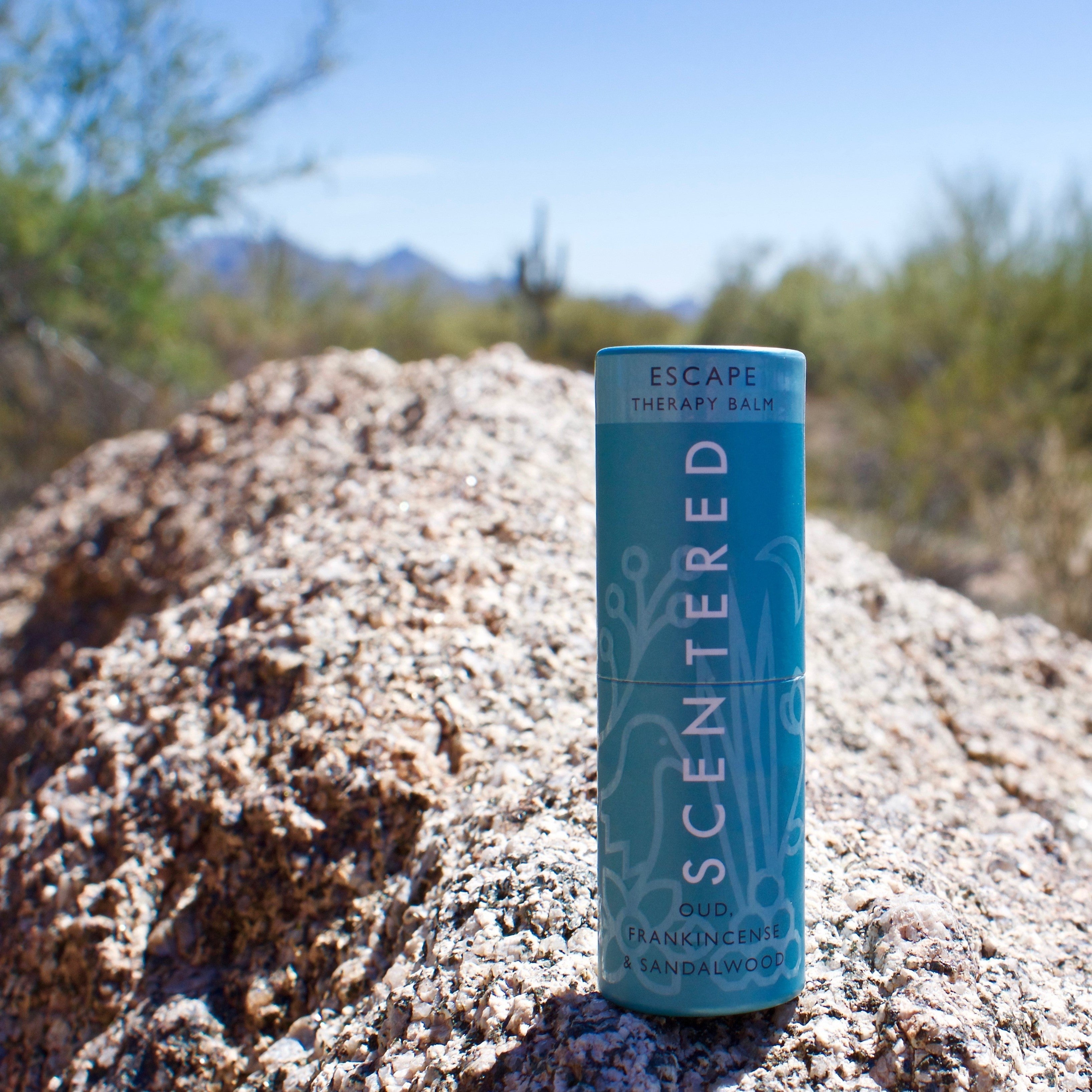 Scentered_Escape_balm_placed_on_a_rock_with_blue_sky_and_bushes_in_background