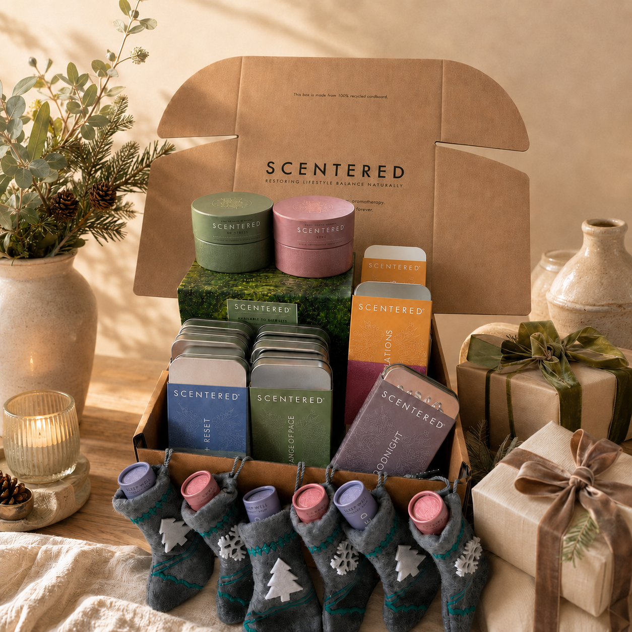 A Scentered by Scentered gift box arrangement on a table, featuring multiple Scentered products including tins, boxes, and Christmas socks, styled with a candle, wrapped gift, and decorative vase in a warm indoor setting.