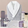 Gray bathrobe with white hood, sleep mask, sleep balm, body oil, candle and various skincare products on a beige background