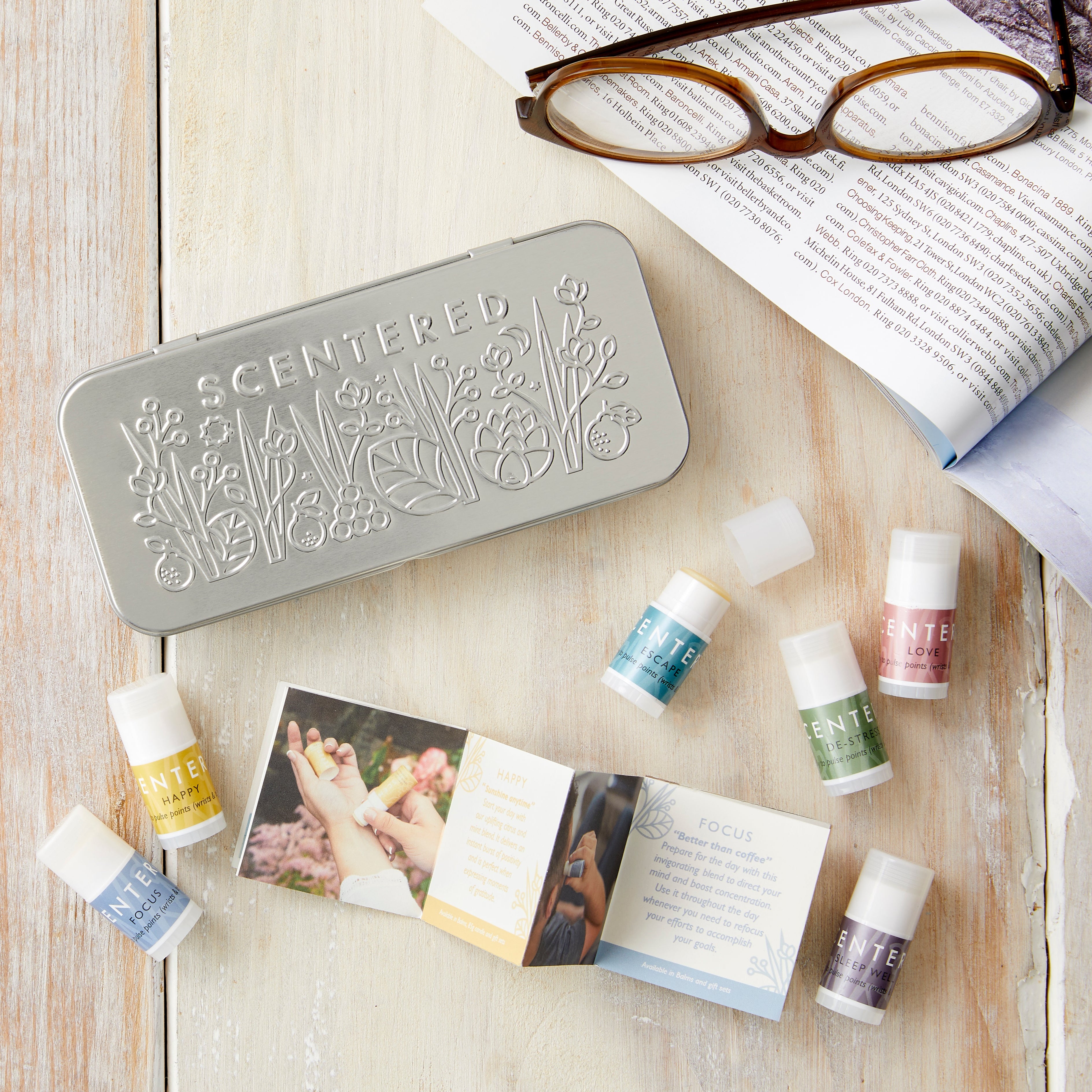 Scentered Discovery Tin featuring embossed floral pattern on the lid of the tin.  Next to the tin on a wooden table is a pair of glasses on an open magazine in the tone of relaxation.  There are also 6 Scentered aromatherapy balms shown scattered on the table which are the contents: Focus, Happy, Escape, De-stress, Love and Sleep Well Aromatherapy blends in 1.5g pulse point balm sticks