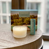A sccentered brand aromatherapy balm and candle set, with the balm next to the candle and a packaged box in the background, all placed on a wooden surface with a window in the background showing a tropical beach scene.