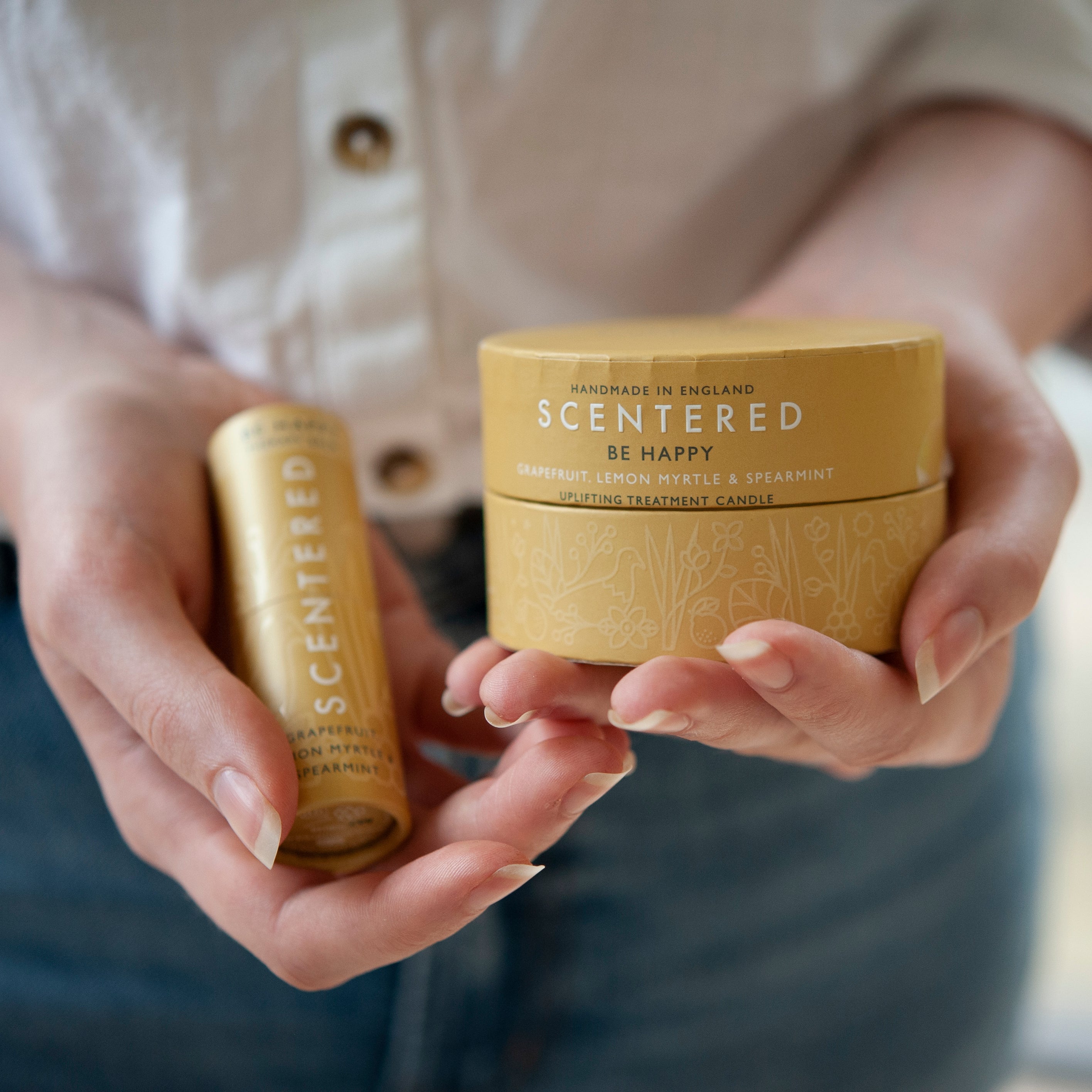 Person holding Scentered Be Happy therapy balm and uplifting treatment candle, featuring grapefruit, lemon myrtle, and spearmint scent in yellow packaging.